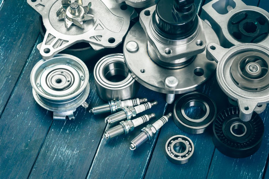 Close-up view of various automotive components including spark plugs, bearings, and metal engine parts arranged on a work surface, representing vehicle maintenance and repair parts.