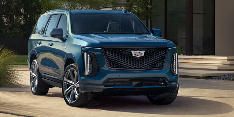 Front three-quarter view of a blue Cadillac Escalade parked outside a modern building, showcasing its grille and LED lighting.
